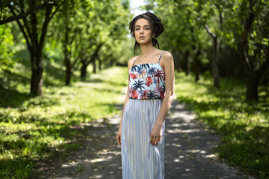 Brunette girl in a dress stands on the nature background. Dress is with silver bottom and flowered top. She holds her hands along the body and looks into the camera with parted lips. Horizontal.