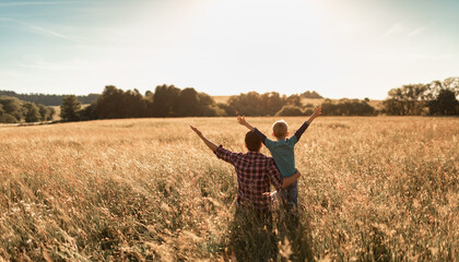 Joyful father son bonding amidst wheat field, arms raised, connecting with nature, under sunny sky.