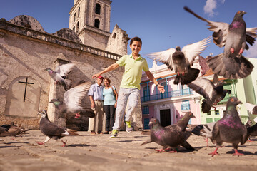 Happy tourist on holidays during vacation trip. Hispanic people traveling in Havana, Cuba. Grandpa and grandson feeding birds, with child running chasing pigeons - obrazy, fototapety, plakaty
