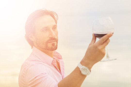Toned picture of sommelier analyzing glass of red wine isolated on blue sky background. Black-haired mature man happy looking at glass.