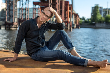 Stylish barefoot guy in glasses sits on the quay on the background of the river and the bridge. He wears blue jeans and a dark shirt. Male looks up. Outdoors. Horizontal.