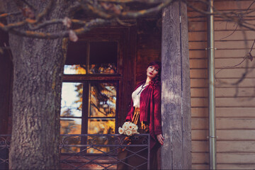 Beautiful young woman with flowers in long skirt and wool red scarf standing on old house balcony. Early fall. Outside shot. Ambient light.