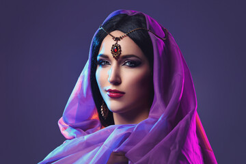 Naklejka premium Beautiful young caucasian woman with long black hair and arabian style makeup. Vintage accessory on head. Purple fabrics. Studio shot over dark background.