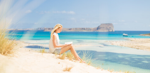 Relaxed woman enjoying sun, freedom and good book an beautiful sandy beach of Balos in Greece. Young lady reading, feeling free and relaxed. Vacations, freedom, happiness, enjoyment and well being.