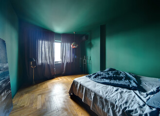 Bedroom in a modern style with turquoise walls and a parquet on the floor. There is a bed with blue pillow and blanket, blue picture, lamps, stand with candlestick, window with curtains, conditioner.