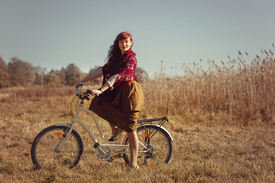 Beautiful young woman in long skirt and wool scarf riding retro style bicycle in field. Outside shot. Autumn season. Copy space.