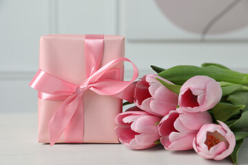 Beautiful gift box with bow and pink tulips on white table, closeup
