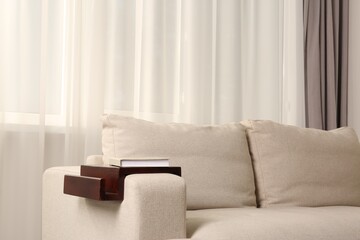 Wooden armrest table with book on sofa in room. Interior element