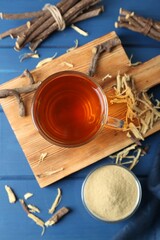 Aromatic licorice tea in cup, dried sticks of licorice root and powder on blue wooden table, flat lay