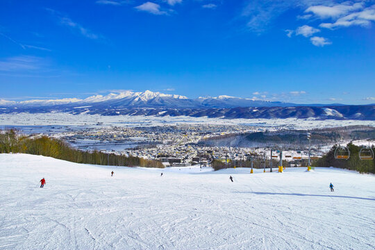 快晴の富良野スキー場から見る十勝岳連峰、北の峰ゾーンの広大な初心者コースを滑るスキーヤー達
