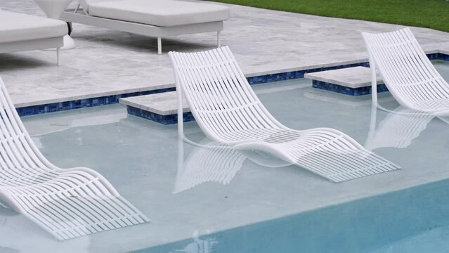 Exquisite exterior of lounge area near swimming pool with sun loungers. Three white deck chairs of curved shape placed in transparent water