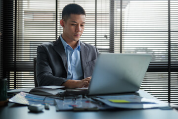 Young asian businessman sitting in bright modern workplace and working with laptop computer.