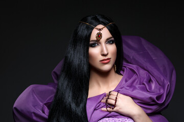 Beautiful young caucasian woman with long black hair and arabian style makeup. Vintage accessory on head. Purple fabrics. Studio shot over dark background. Copy space.