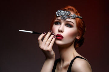 Beautiful young woman with smoky eyes and full red lips holding cigarette holder. Massive crystal hair accessory on head. Retro styling. Studio beauty shot. Copy space.