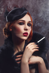 Beautiful young woman with smoky eyes and full red lips holding cigarette holder. Vintage head piece. Retro styling. Studio beauty shot over smoky background. Copy space.
