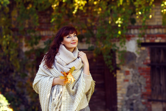 Beautiful young woman with red hair in wool clothes holding yellow autumn leaves. Outdoor fall shot. Natural background. Copy space.