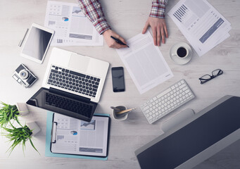 Businessman working at office desk and signing a document, computers and paperwork all around, top view