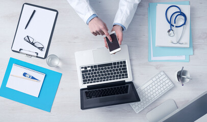 Doctor working at office desk and using a mobile touch screen phone, computer and medical equipment all around, top view