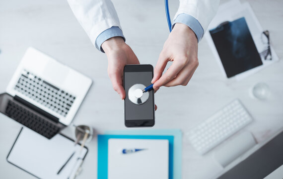 Doctor checking heartbeat on mobile phone using a stethoscope, desktop with medical equipment on background, medical app concept
