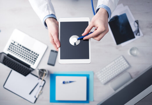Doctor checking heartbeat on a digital tablet using a stethoscope, desktop with medical equipment on background, top view