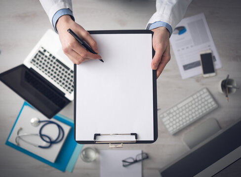 Doctor writing medical records on a blank sheet and holding a clipboard, medical equipment and desktop on background, top view