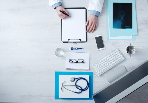 Professional doctor writing medical records on a clipboard with computer and medical equipment all around, desktop top view with copyspace