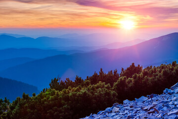 Colorful sunset or sunrise with sunshine and clouds above blue misty mountain silhouettes. Fir bushes and stones on the hill side. Horizon skyline panorama landscape with mountains. - Powered by Adobe
