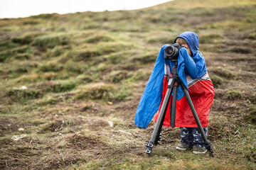 Little kid in a multi-colored big size windcheater is making a photo on the background of mountains. He is looking into the viewfinder of the camera which is on the tripod. Outdoors. Horizontal.