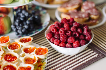 Table with snacks. In the foreground a dish with raspberries and tarts with red caviar.