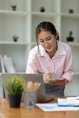 Office worker or businesswoman, attractive Asian woman in eyeglasses researching Analyze information about the business plan. take notes in a book Notebook while using a laptop computer at her desk.