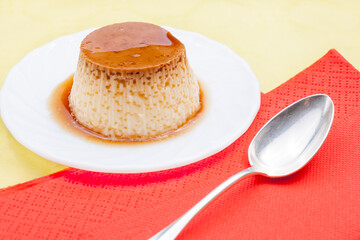 Closeup of vanilla flan with caramel red napkins and spoon on yellow table