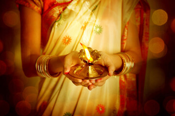 Close up Indian woman in traditional sari lighting oil lamp and celebrating Diwali or deepavali, fesitval of lights at temple. Female hands holding oil lamp.