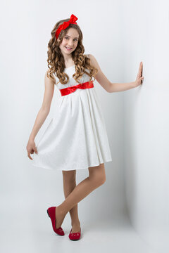 Beautiful teenage girl with long curly hair and red ribbon bow on head wearing white dress standing near wall. Happy expression. Studio portrait on white background. Copy space.