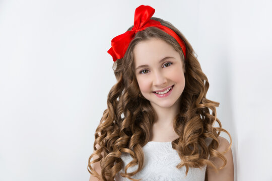 Beautiful teenage girl with long curly hair and red ribbon bow on head wearing white dress. Happy expression. Studio portrait on white background. Copy space.