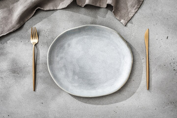 Minimal place setting in gray tones. Gray plate, gold cutlery and linen napkin on concrete table...