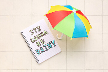 Notebook with text IT'S GONNA BE RAINY and umbrella on white tile background. Weather forecast...