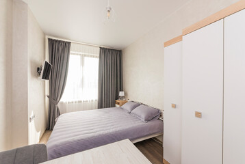 Interior design, bedroom with one window and a large double bed near a white wardrobe