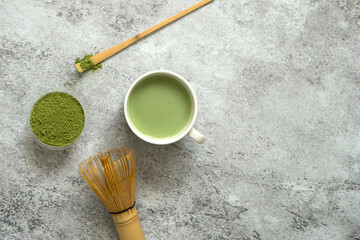Matcha tea in a white cup with bamboo whisk and spoon on a gray concrete background. This powdered...