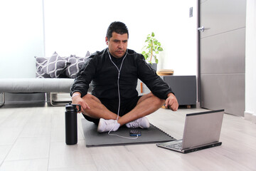 Dark-haired 40-year-old Latino man practices yoga in his living room with an online instructor for...