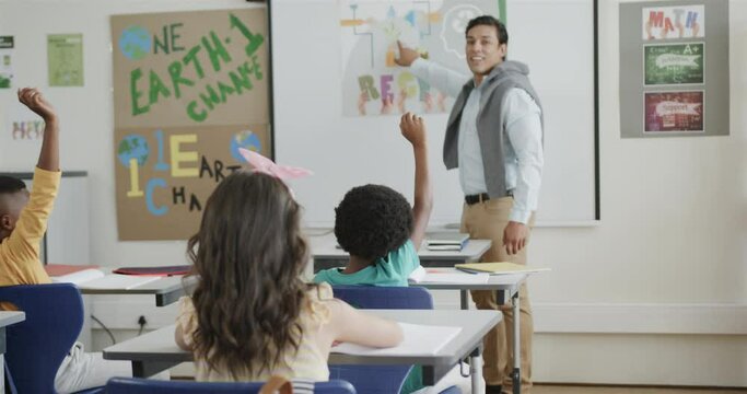 Diverse male teacher and children raising hands at desks in elementary school class, slow motion