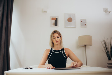 Obraz premium Start of a work day. Young woman opening laptop, getting ready for work in the morning at the desk near windows in a beautiful office.