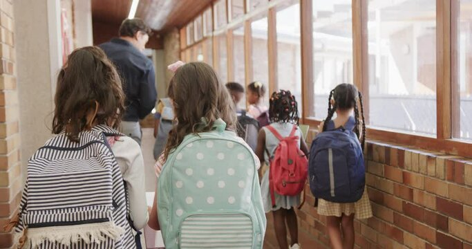 Happy diverse male teacher and children with bags walking in elementary school corridor, slow motion