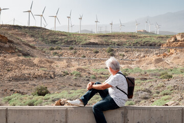 Active senior man sitting outdoor in a trekking day looking at windmill farm on the mountain. Elderly white-haired grandfather enjoy vacation or retirement appreciating sustainable energy