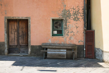Fototapeta na wymiar Cinque Terre, Italy - rugged building exterior with peeling paint, worn wooden double doors, an old wood bench, used shutter to the side. House in a state of disrepair. Home repair service background.