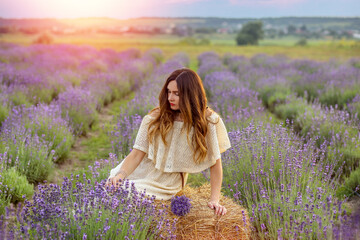 Caucasian girl in a light dress sitting on a haystack in a lavender field. Provence, France. Summer and vacation concept.