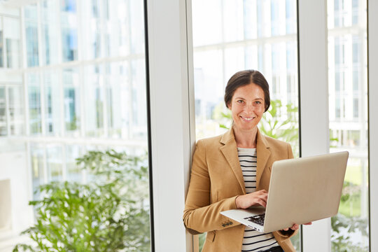 Portrait of happy female entrepreneur holding laptop leaning on window in office lounge