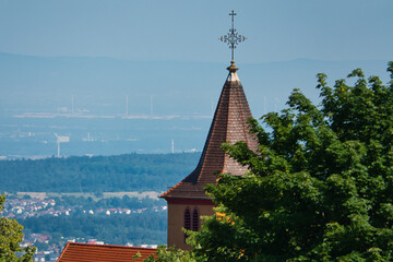 Eine Kirchturmspitze mit einem Kreuz aus Metall auf dem Dach, halb verdeckt von einem Baum, mit...
