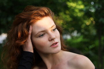 Portrait of sensual ginger girl with long red hair and freckles standing near the tree in evening sunlight. Young seductive model, natural female beauty and desire