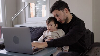 A young man trying to work on laptop holding his son at home. Remote work