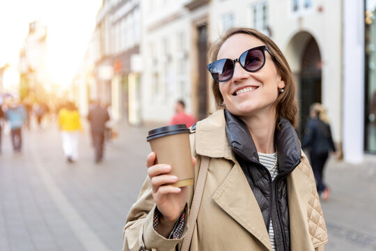 Portrait young beautiful happy smiling woman drinking coffee to go tea in eco sustainable paper cup wear biege trench coat enjoy walking european city street. Stylish female person urban lifestyle